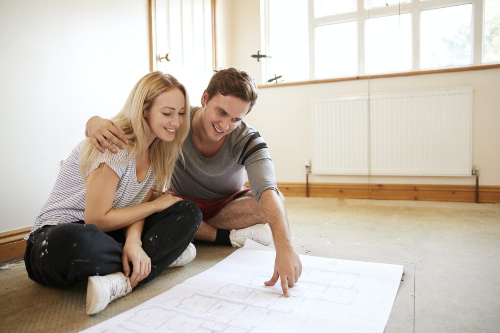 Couple Sitting On Floor Looking At Plans In Empty Room Of New Home